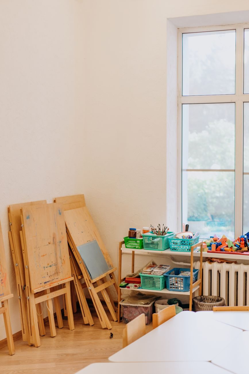 An art corner with folding wooden easels against a white wall, baskets of brushes and blocks on a shelf, and a tall window onto trees.