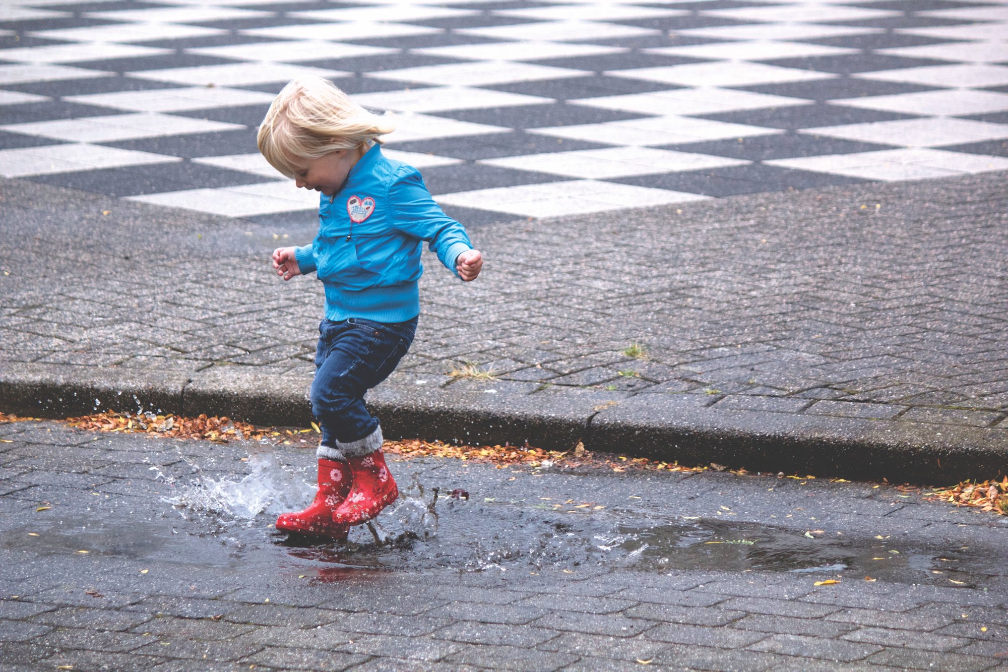 A toddler in a blue jacket and red boots mid-jump over a puddle on a cobbled path.