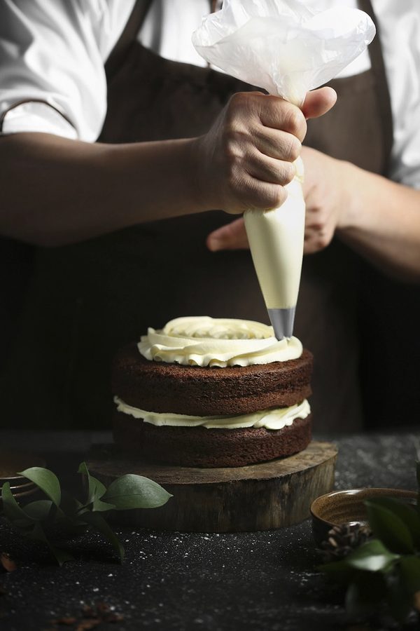 Dana finishing a wedding tier — piping cream onto a two-tier chocolate cake.