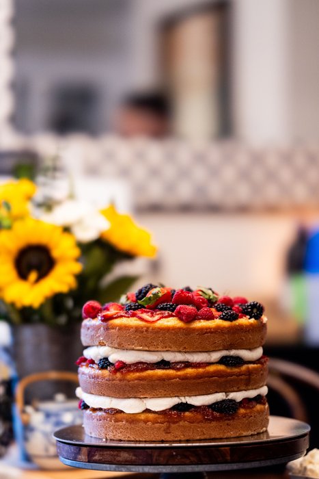 A naked layer cake with berries on a stand in the front window, sunflowers behind it.