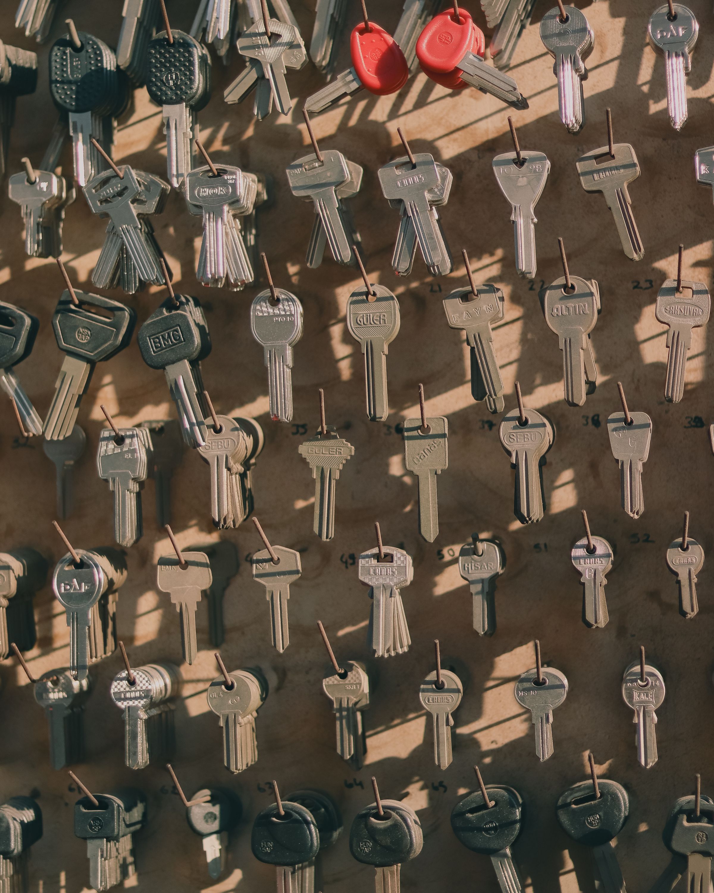 A locksmith&rsquo;s key board: dozens of cut keys and blanks on numbered hooks over tan pegboard, two red-headed car keys near the top.