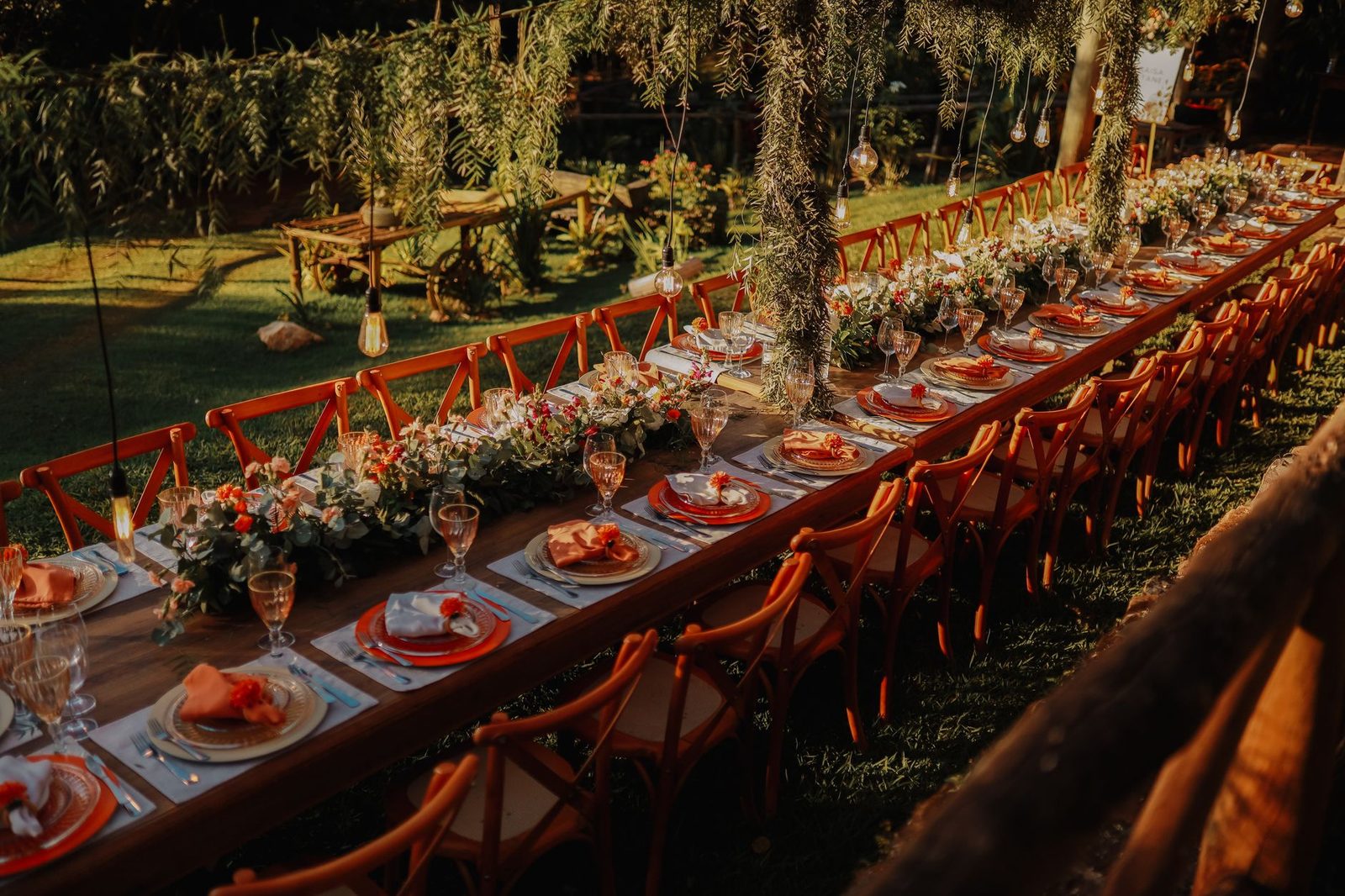 A long wooden table set for a party in a garden at golden hour, terracotta cross-back chairs down both sides and a runner of greenery and flowers along the middle