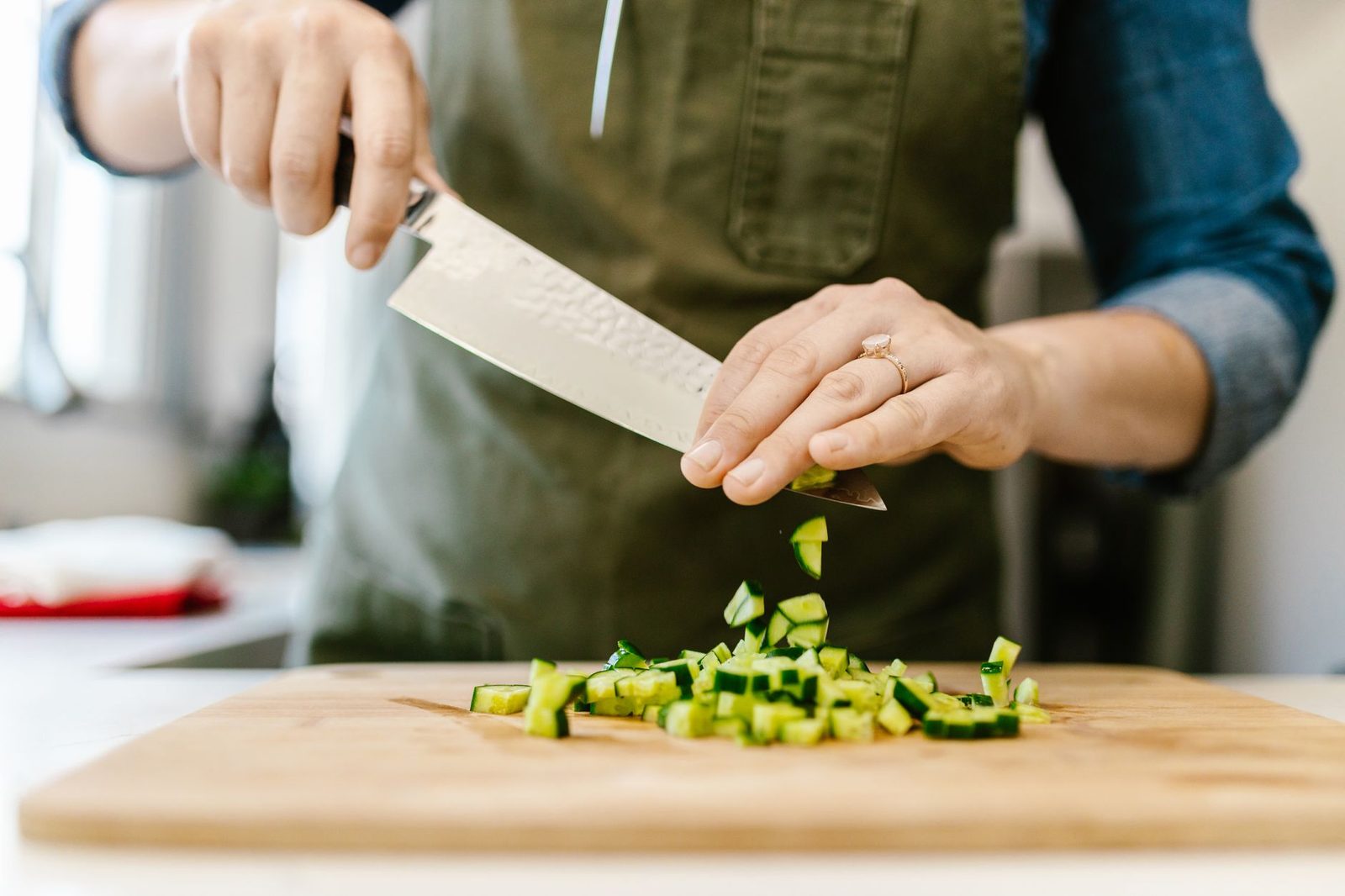 Hands in an olive apron slicing cucumber into even rounds on a wooden board in a bright kitchen