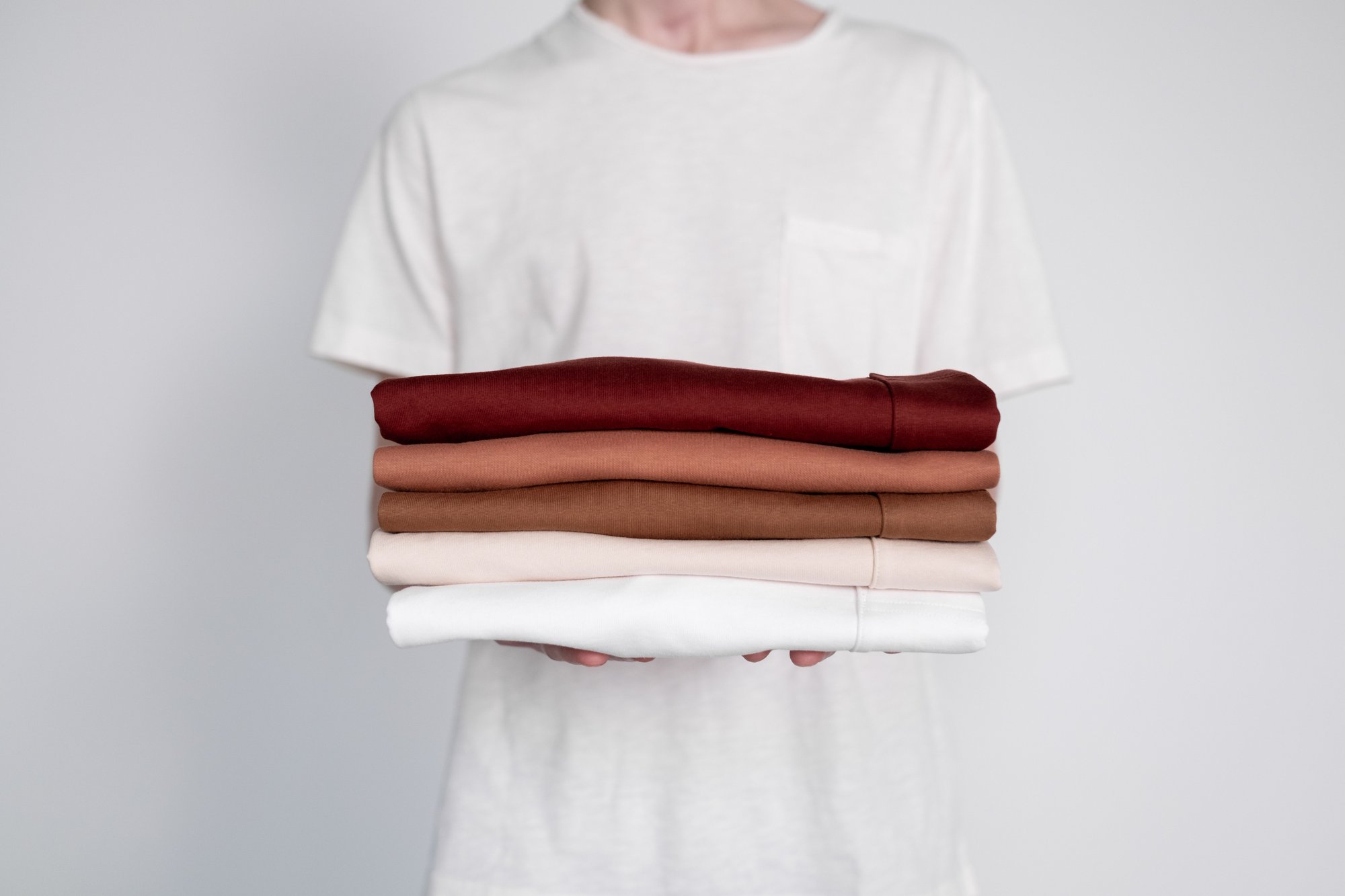 A stack of folded shirts in neutral colors held in two hands.