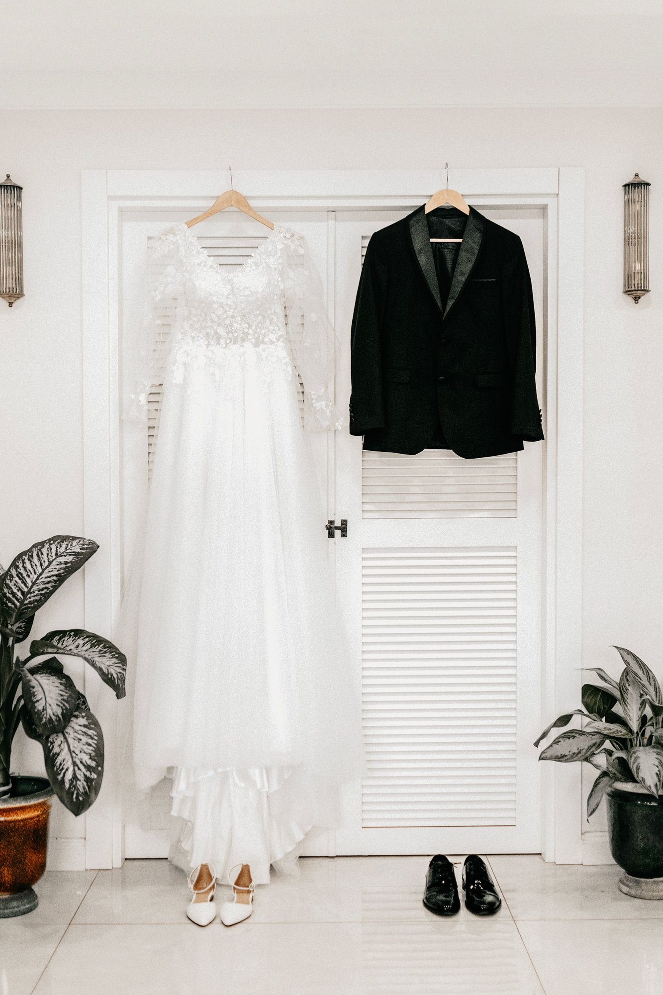 A white wedding gown on a hanger beside a dark suit, in a bright room.
