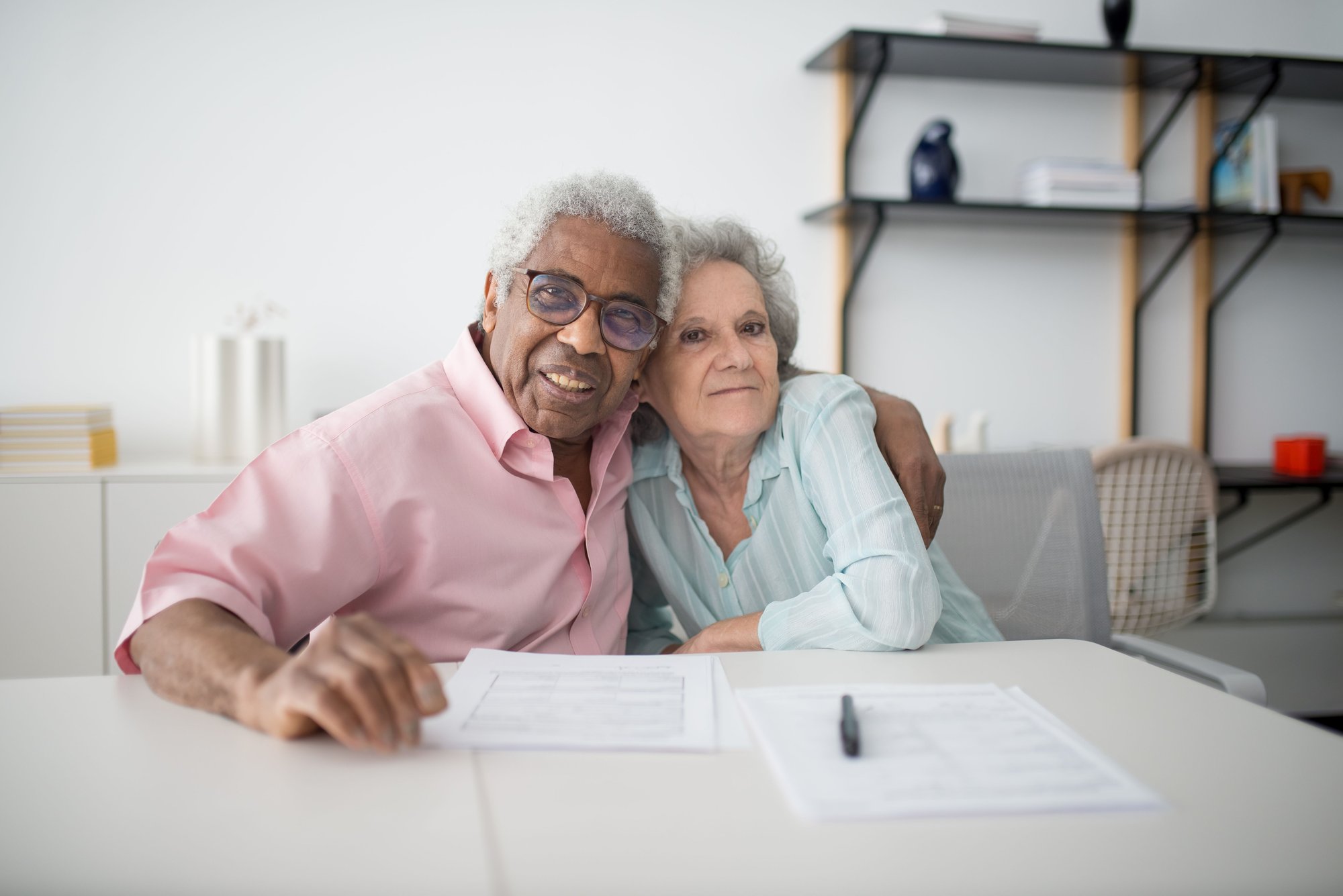 An older couple at a table with paperwork, his arm around her shoulders, both smiling.