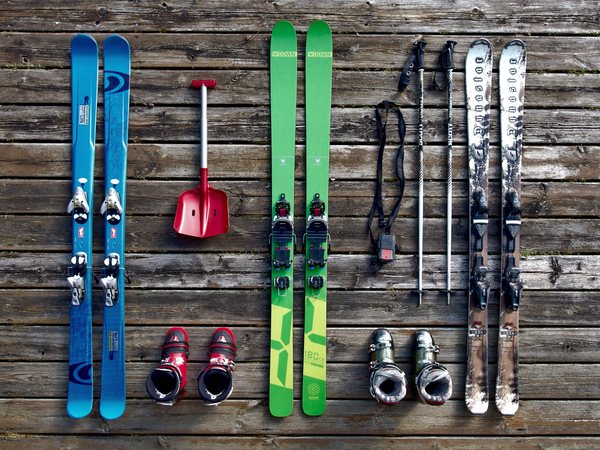 A full ski package laid out on weathered boards: two pairs of skis, boots, poles and a red shovel.