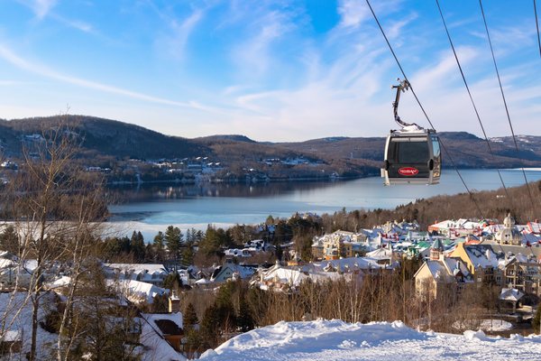 A gondola cabin riding its cable above a snow-covered ski village, with a frozen lake and low hills behind.