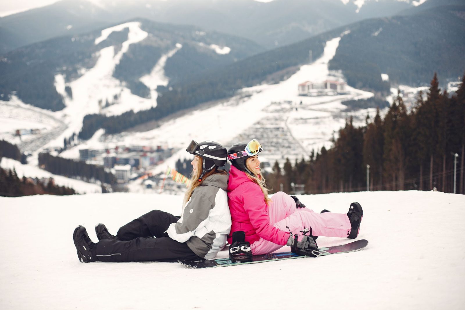 Two snowboarders in pink and grey snow gear sitting back-to-back on their boards, with a snowed-in resort town in the valley behind them.
