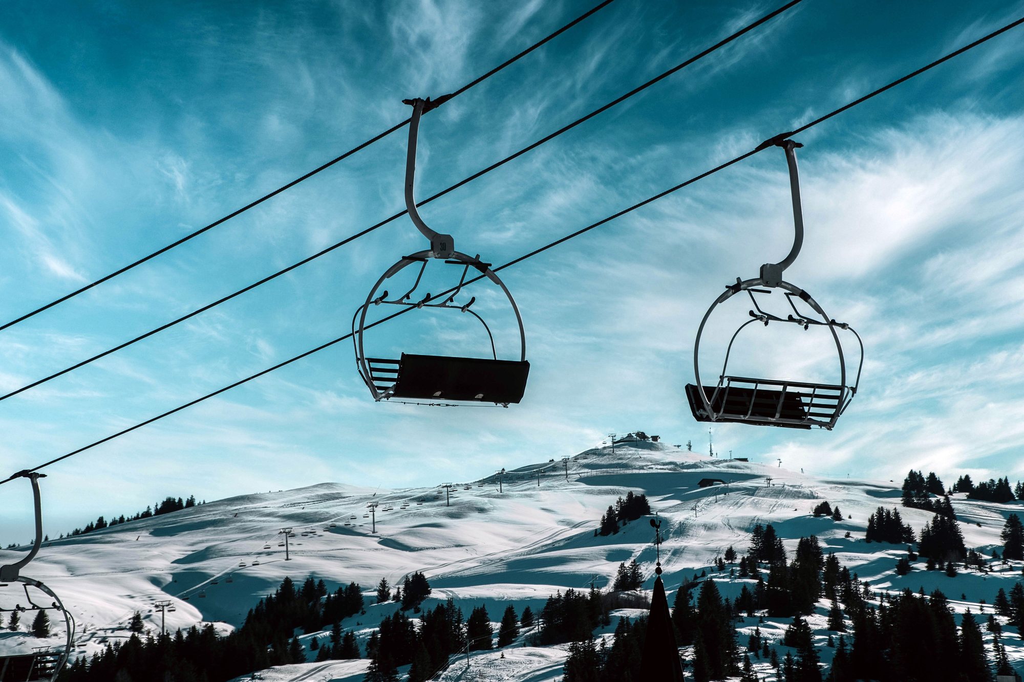 Two empty chairlift seats hanging against a bright blue sky, groomed runs and scattered pines on the mountain below.