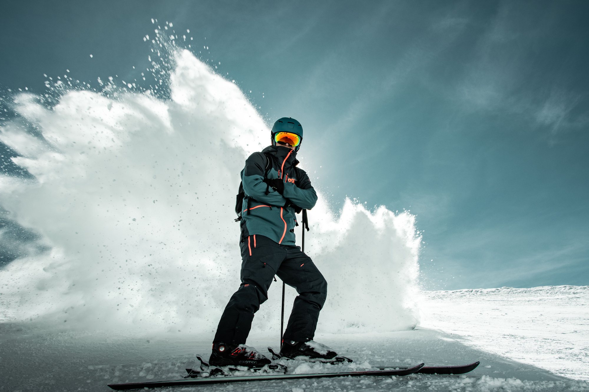 A skier in a teal jacket stopping hard on the slope, a wall of powder thrown up against a deep blue sky.