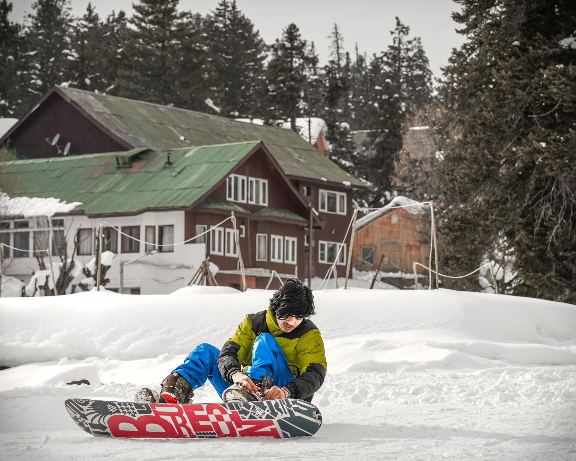 A snowboarder in a yellow jacket sitting in the snow strapping into their board, wooden lodges and pines behind.