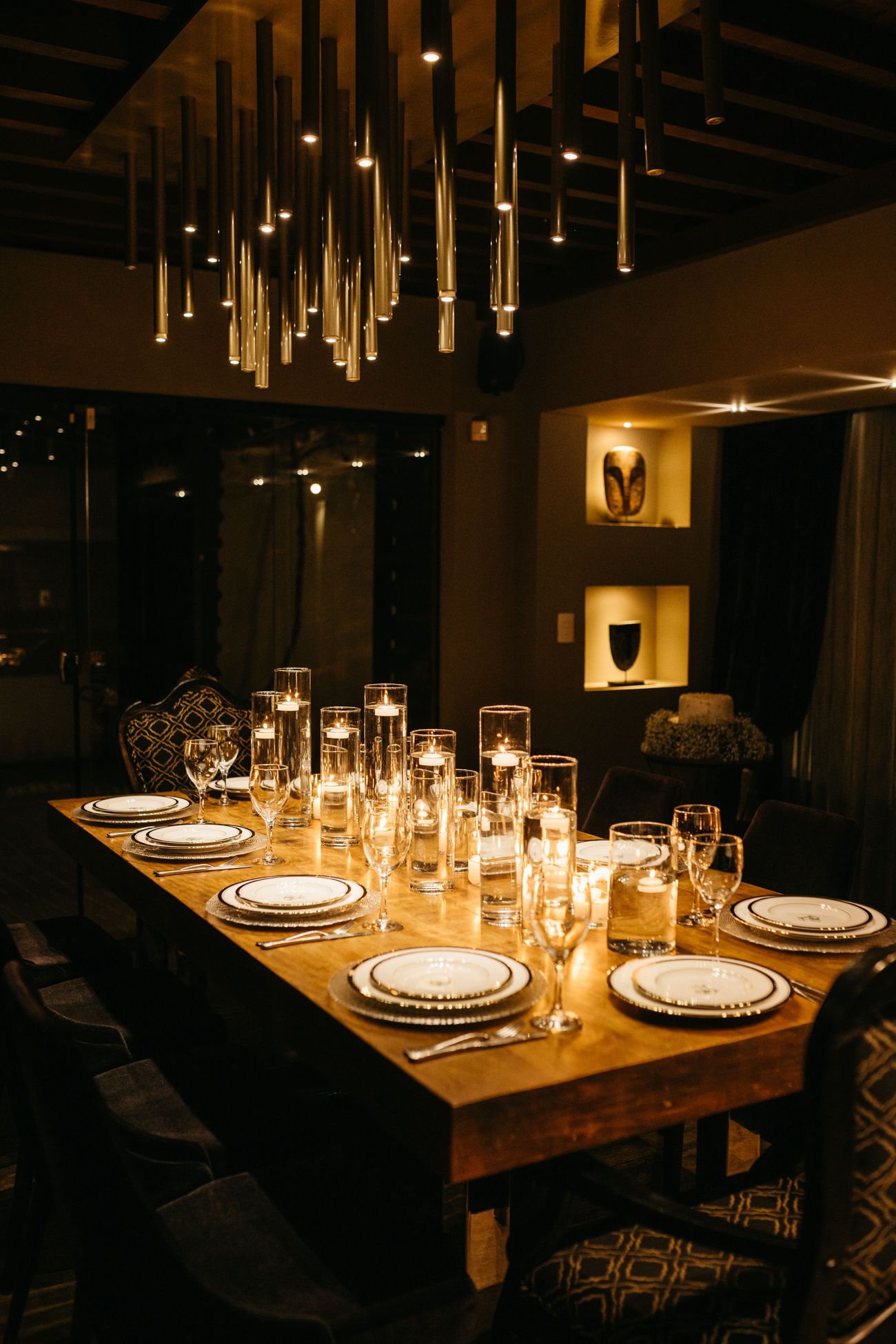 A long table laid with candles in tall glass holders in a dark room, slender pendant lights hanging above it.