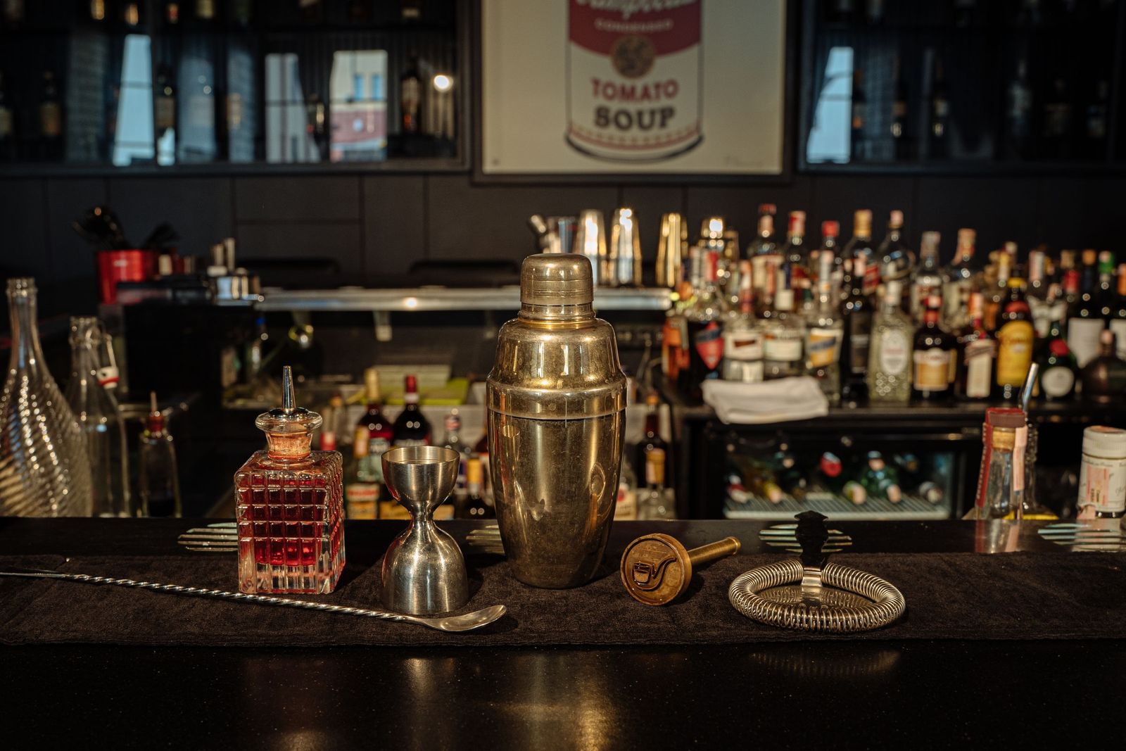 A bar counter laid out with a shaker, jiggers and strainers, bottles ranked behind them